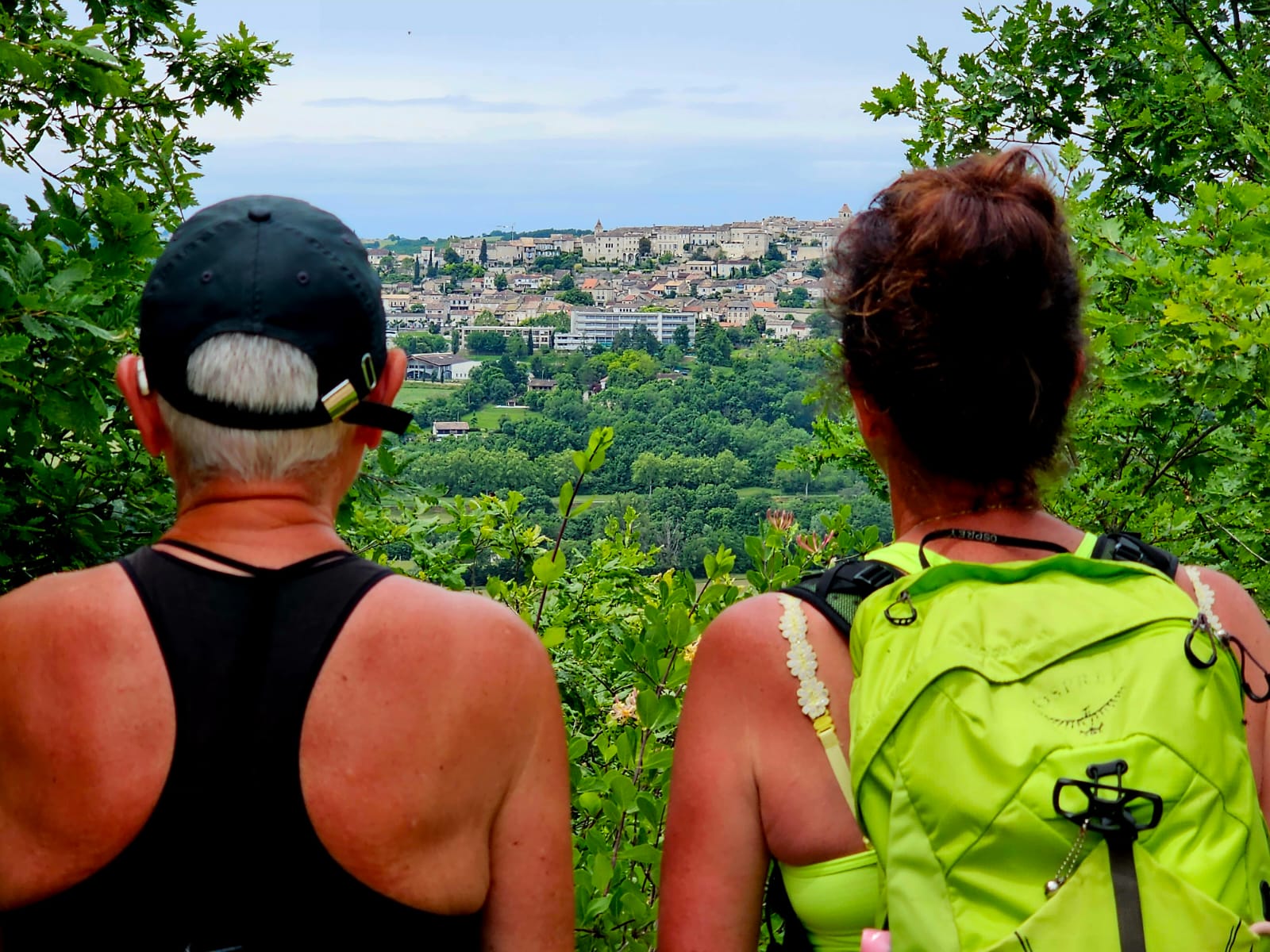 rugaanzicht van 2 wandelaarsters kijkend op een dorp in de verte met veel groen op de via podiensis